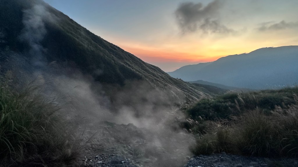 一幅山脈與夕陽交織的風景，山坡上冒出蒸汽，周圍草木茂盛，呈現出自然的壯麗與寧靜。